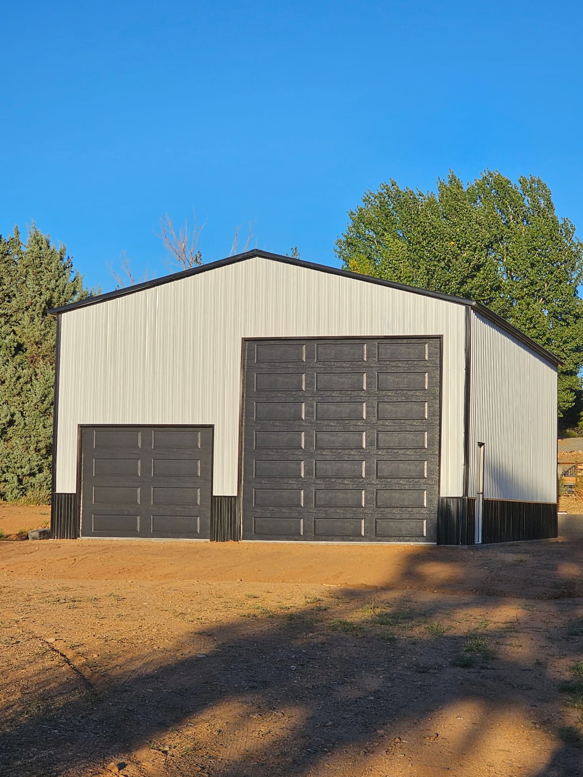 Light gray steel garage with black trim and wainscot and two dark roll-up doors, one extra tall, on a dirt lot