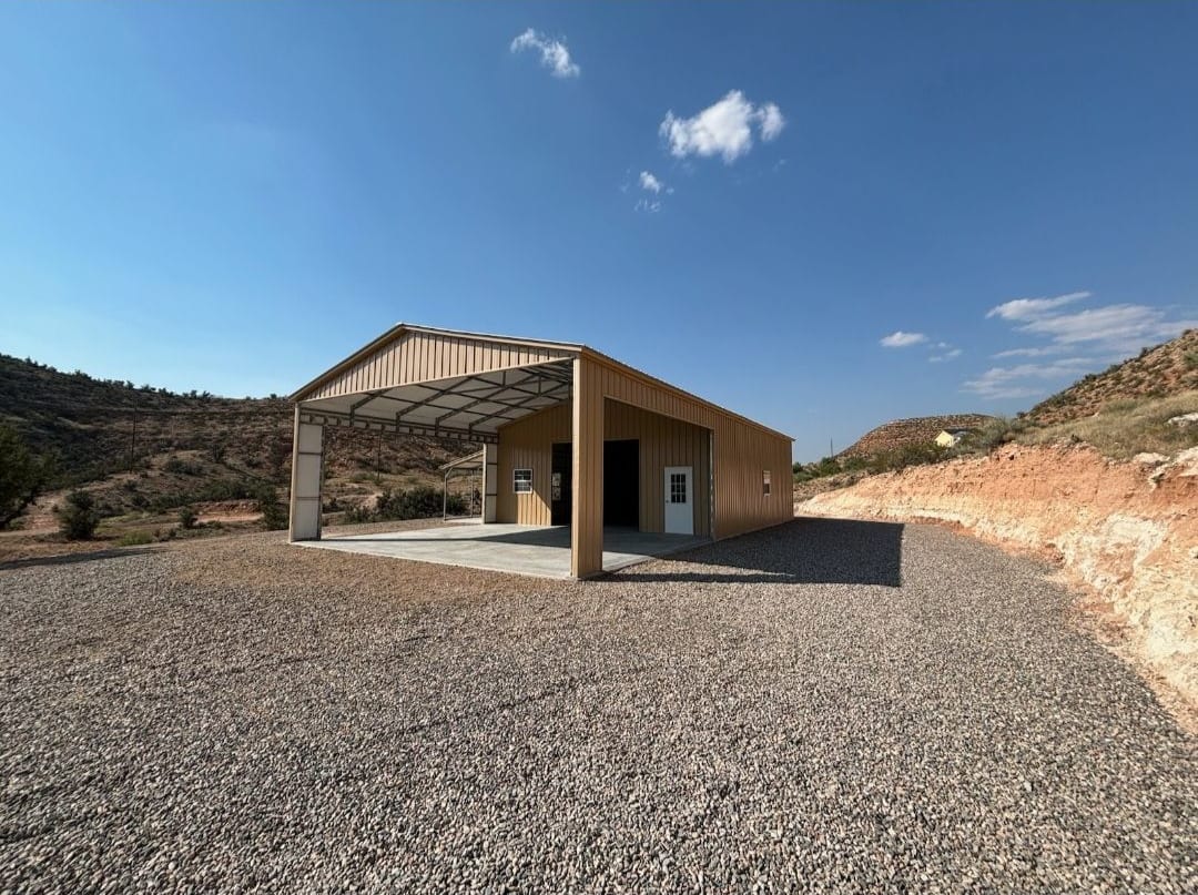 Tan metal building with an open covered bay in front of an enclosed room with a walk-in door, on gravel